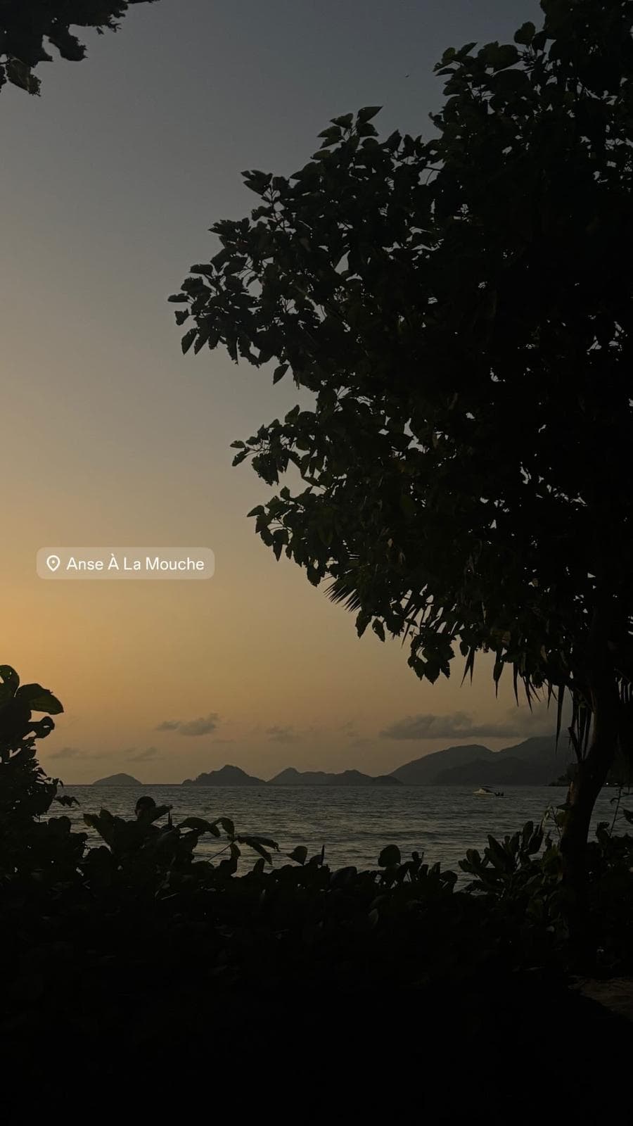 Sunset over the water at Anse À La Mouche, headlands silhouetted on the horizon behind a dark tree canopy.
