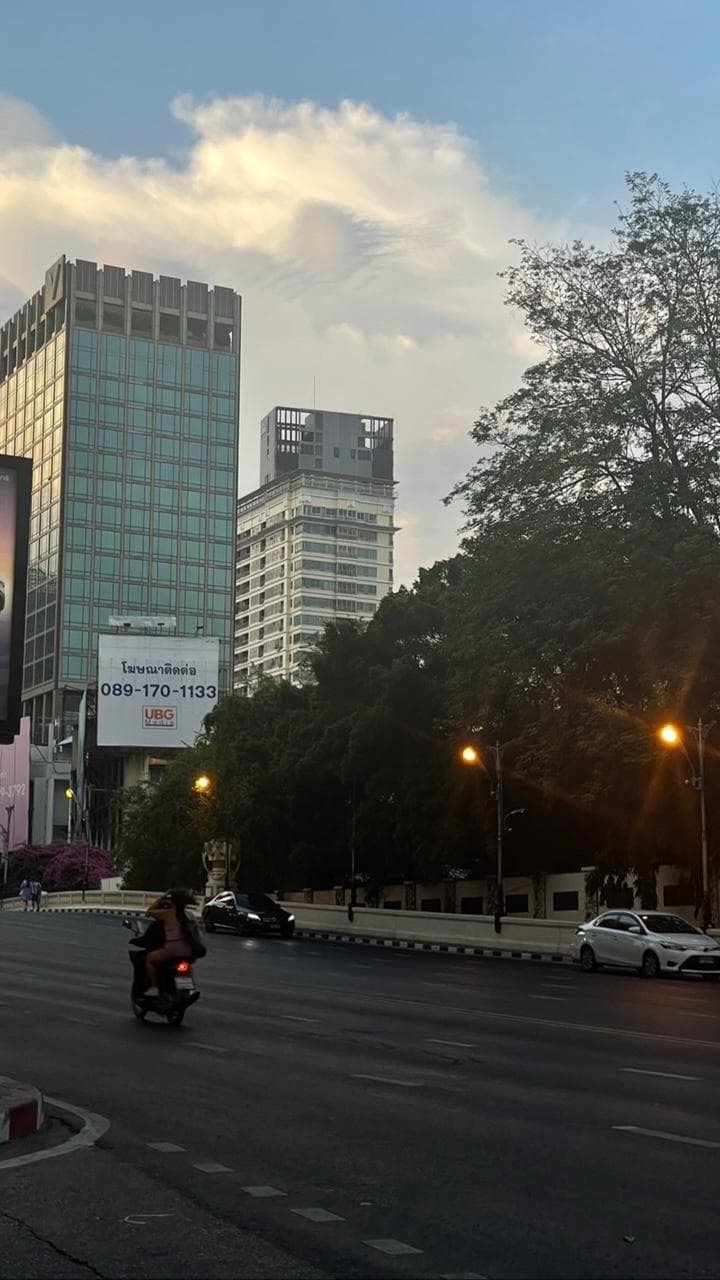 A wide city road at dusk, tower blocks catching the last light above the treeline.